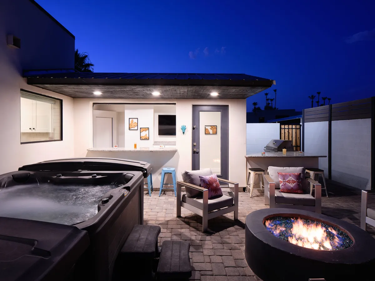 A cozy, inviting backyard space that includes a bubbling hot tub, a warm fire pit surrounded by outdoor seating, and an outdoor kitchen/bar area. The space is designed for socializing, stargazing, and enjoying meals or drinks al fresco, all in a private, landscaped setting.