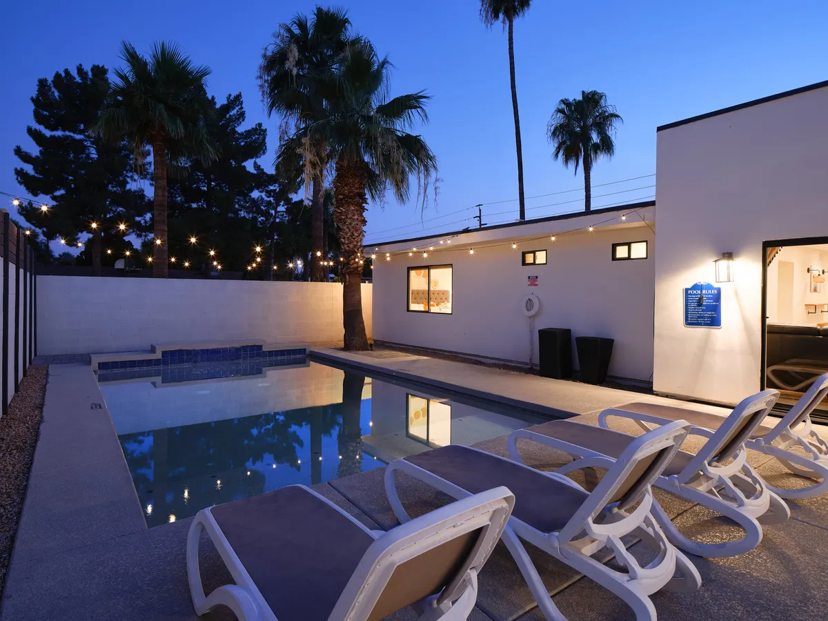 A beautifully landscaped backyard featuring a sparkling swimming pool surrounded by comfortable lounge chairs, string lights stretched overhead, palm trees swaying in the background, and an exterior wall that adds privacy and a resort-like feel. The scene is set at dusk, creating a relaxing and festive ambiance perfect for gatherings.