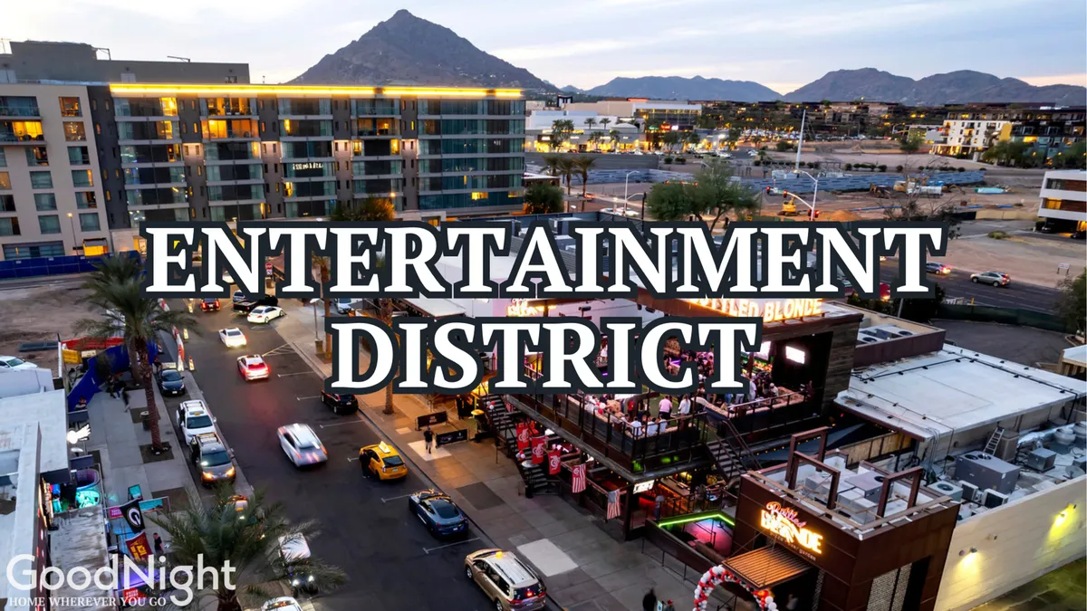 A lively city street lined with entertainment venues, illuminated by city lights with a dramatic mountain backdrop. People and cars animate the street, evoking the energetic nightlife and social scene that define downtown Scottsdale.