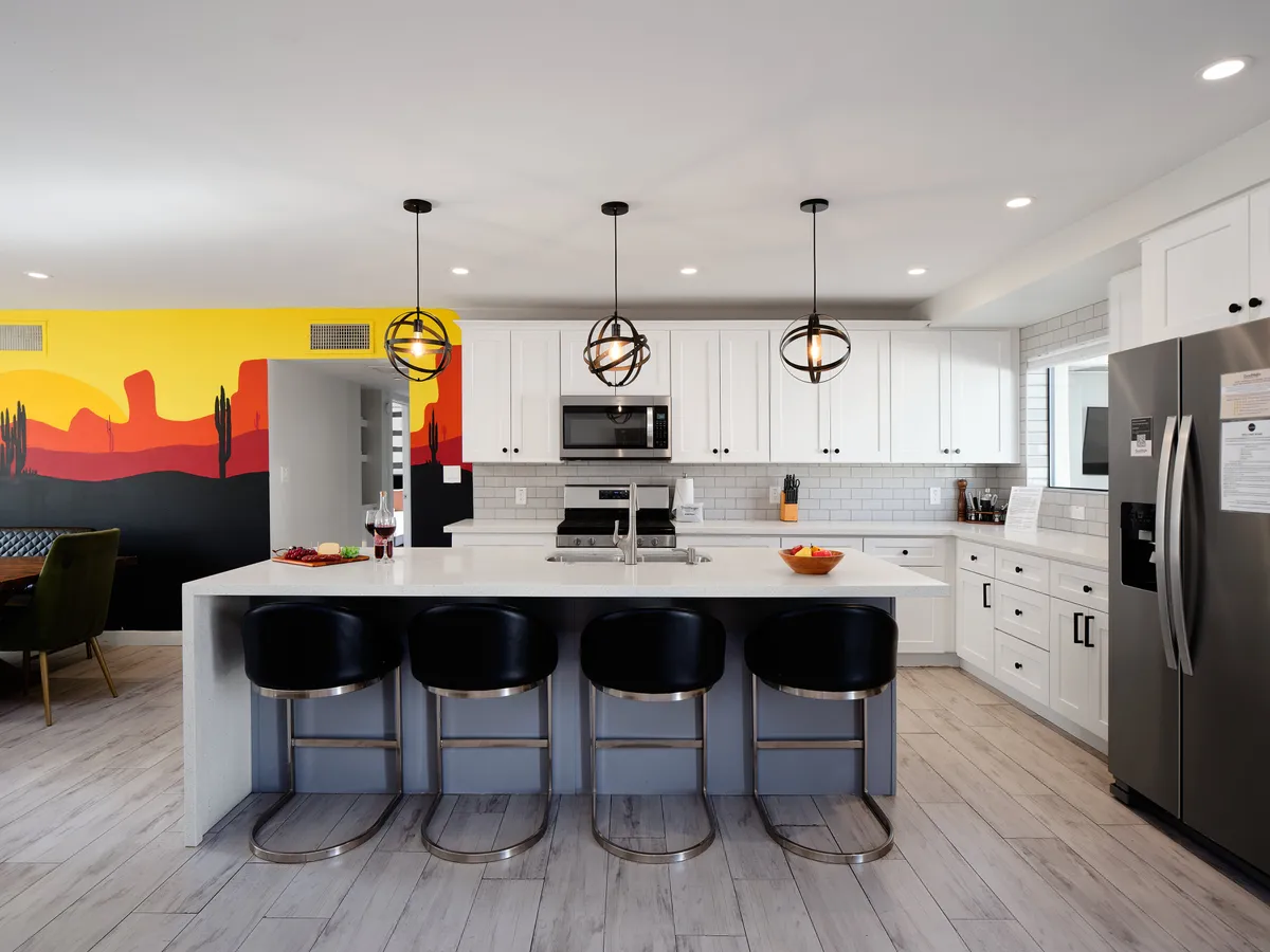 The image spotlights the modern kitchen island with bar seating, colorful mural, pendant lights, and stainless steel appliances. The layout is designed for both cooking and socializing, with plenty of space for group gatherings and casual conversation.