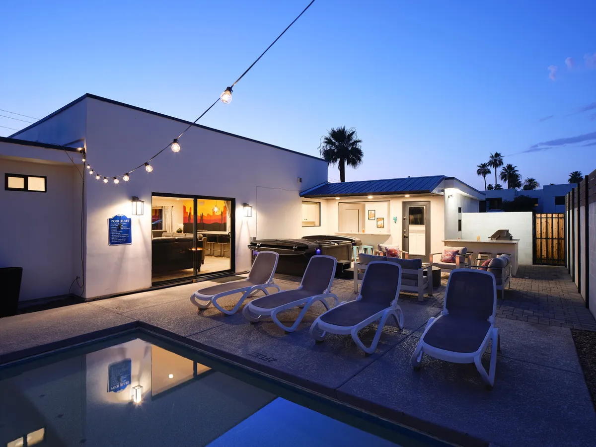 A spacious backyard with a sparkling pool, hot tub, lounge chairs, and string lights overhead. The scene exudes a relaxed, social atmosphere, perfect for groups to unwind and celebrate between outings.