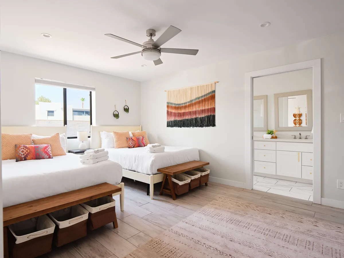 A guest bedroom featuring two beds with decorative pillows, a woven wall hanging, and a mirror reflecting the bathroom, creating a welcoming and flexible sleeping arrangement for families, friends, or guests with children or pets.