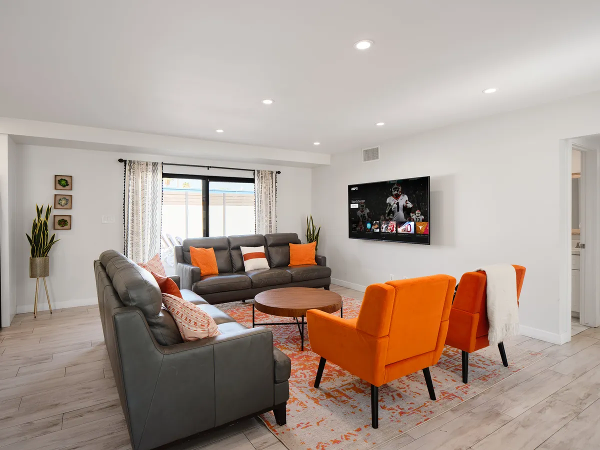 This image focuses on the luminous living room, featuring a plush sofa set, orange accent chairs, a coffee table, and a wall-mounted TV. The space exudes comfort and vibrancy, ideal for unwinding or hosting friends and family after a day of exploration.